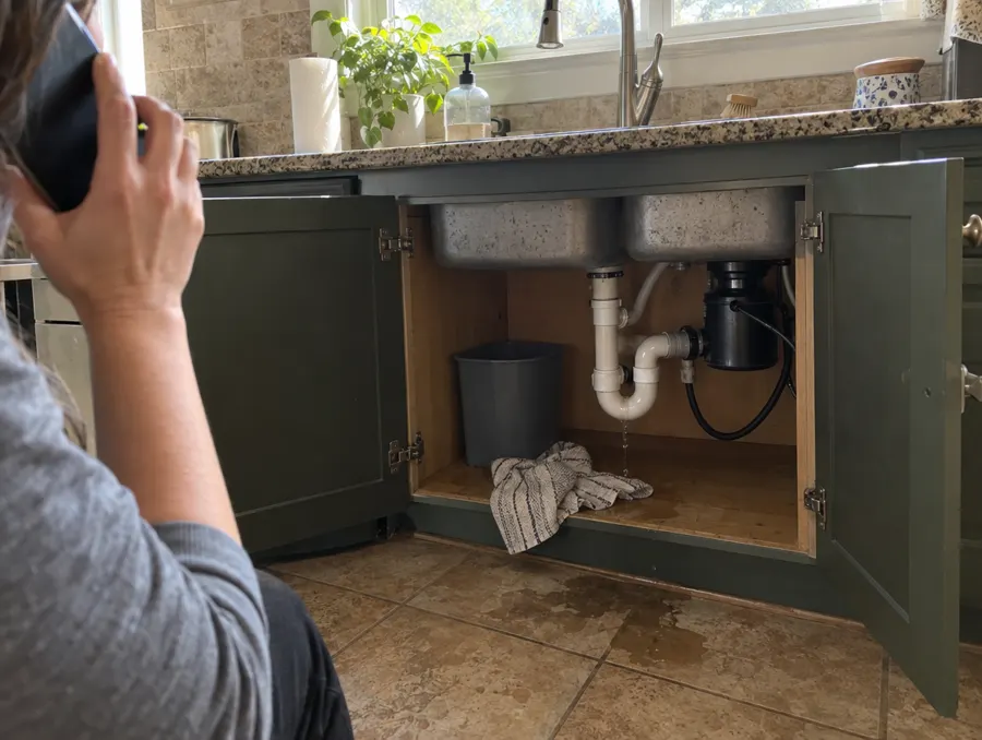 Kitchen sink cabinet open over a wet floor while the homeowner is on the phone