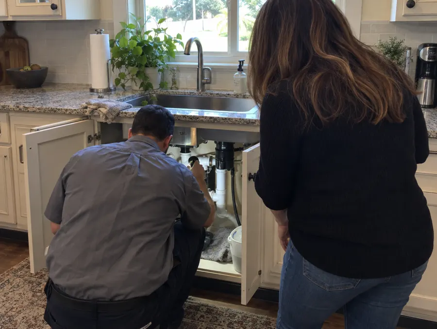 A plumber shines a flashlight under a kitchen sink while the homeowner looks on